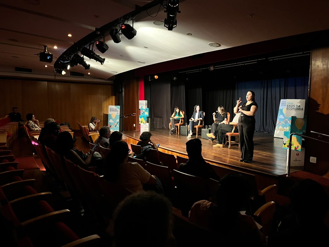 Auditório com plateia acompanhando uma mesa de debate, com intérprete de Libras ao lado do palco.