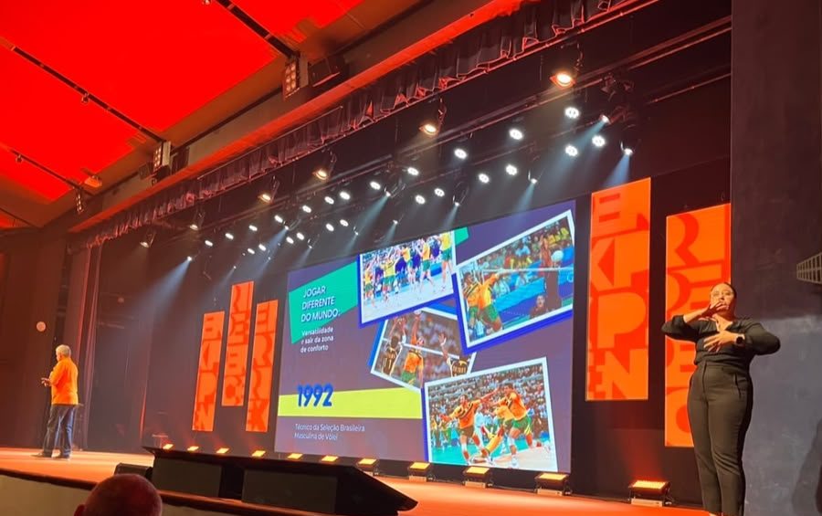 Palco de evento corporativo com palestrante e intérprete de Libras posicionada à direita, diante da plateia.