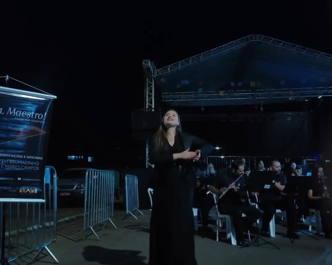 Palco iluminado à noite com orquestra ao fundo e intérprete de Libras posicionada à frente.