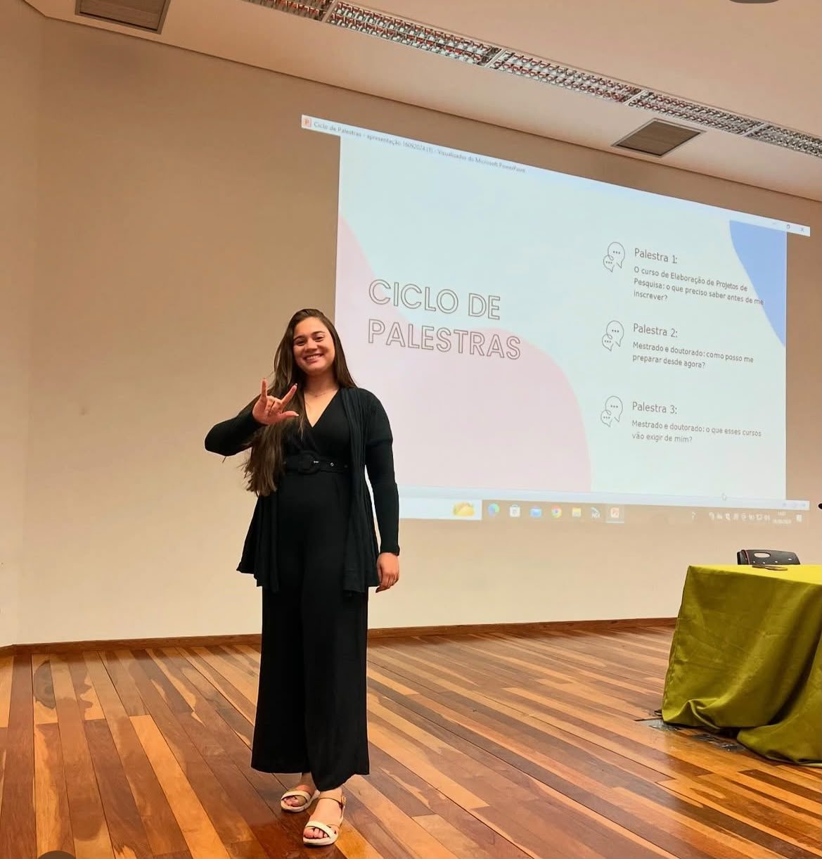 Intérprete de Libras sinalizando no palco de um ciclo de palestras, com a programação do evento projetada ao fundo.